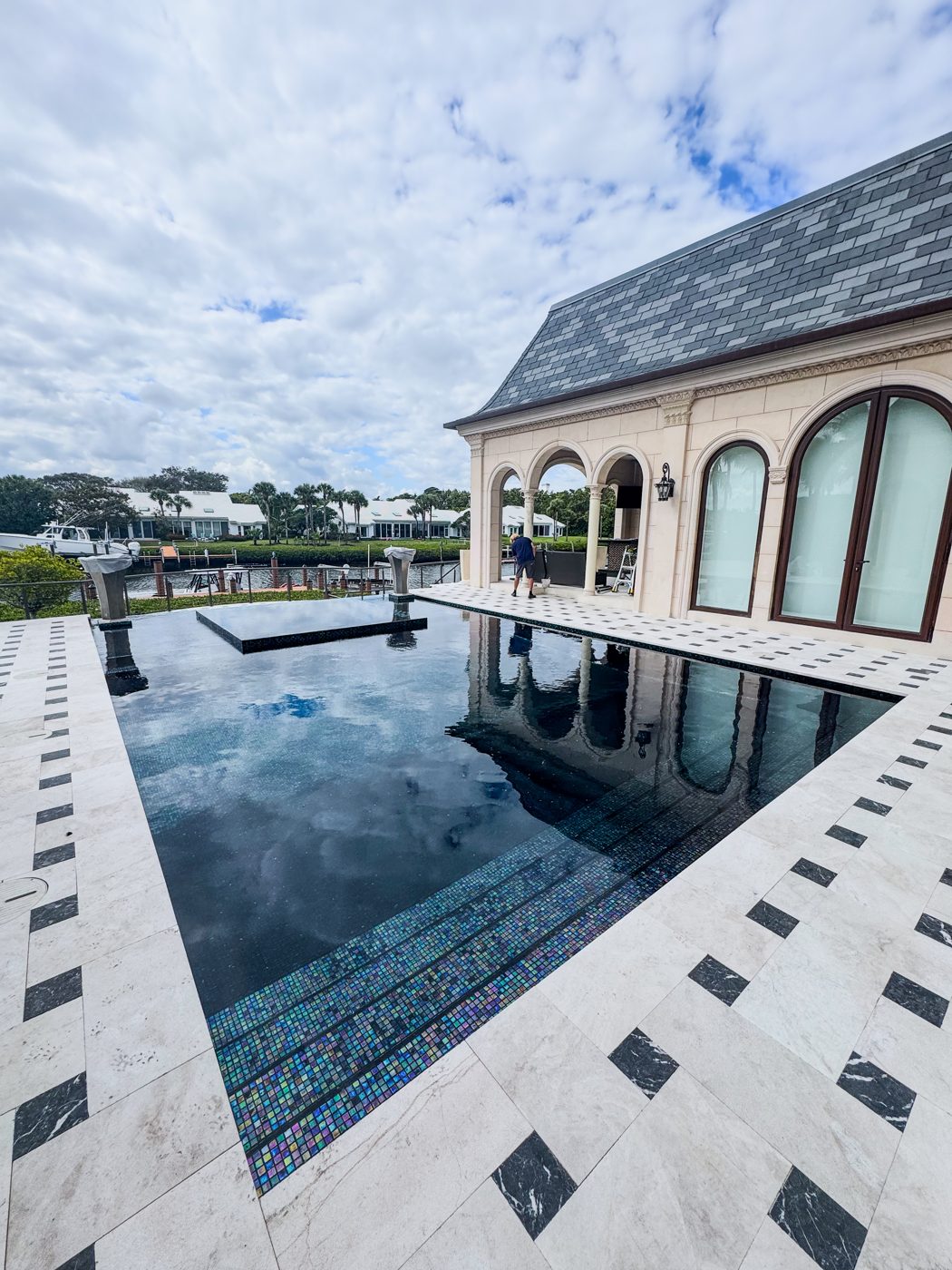 Daytime view across the dark glass-tile pool to the waterway, with the new marble deck and black inlays